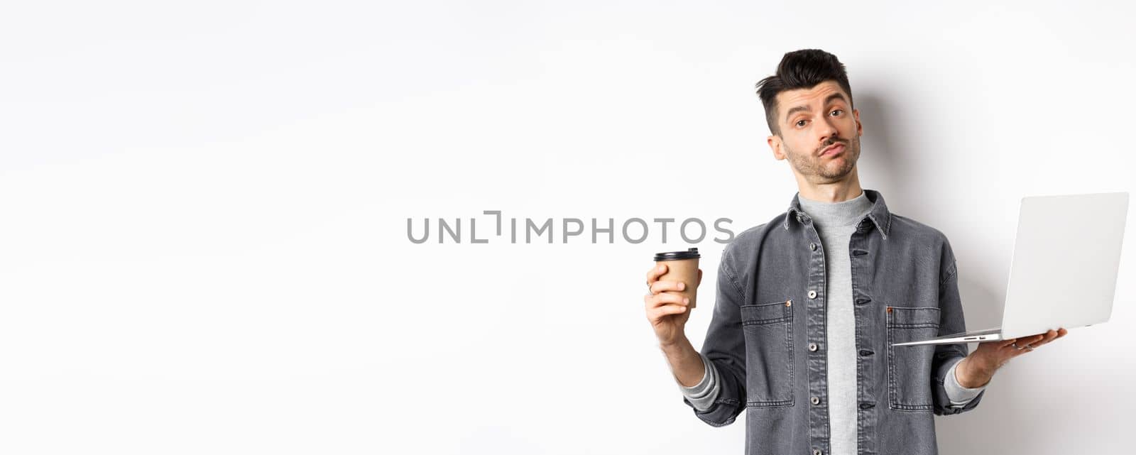 Handsome young man trying new coffee from cafe, holding cup and working on laptop, standing thoughtful on white background.