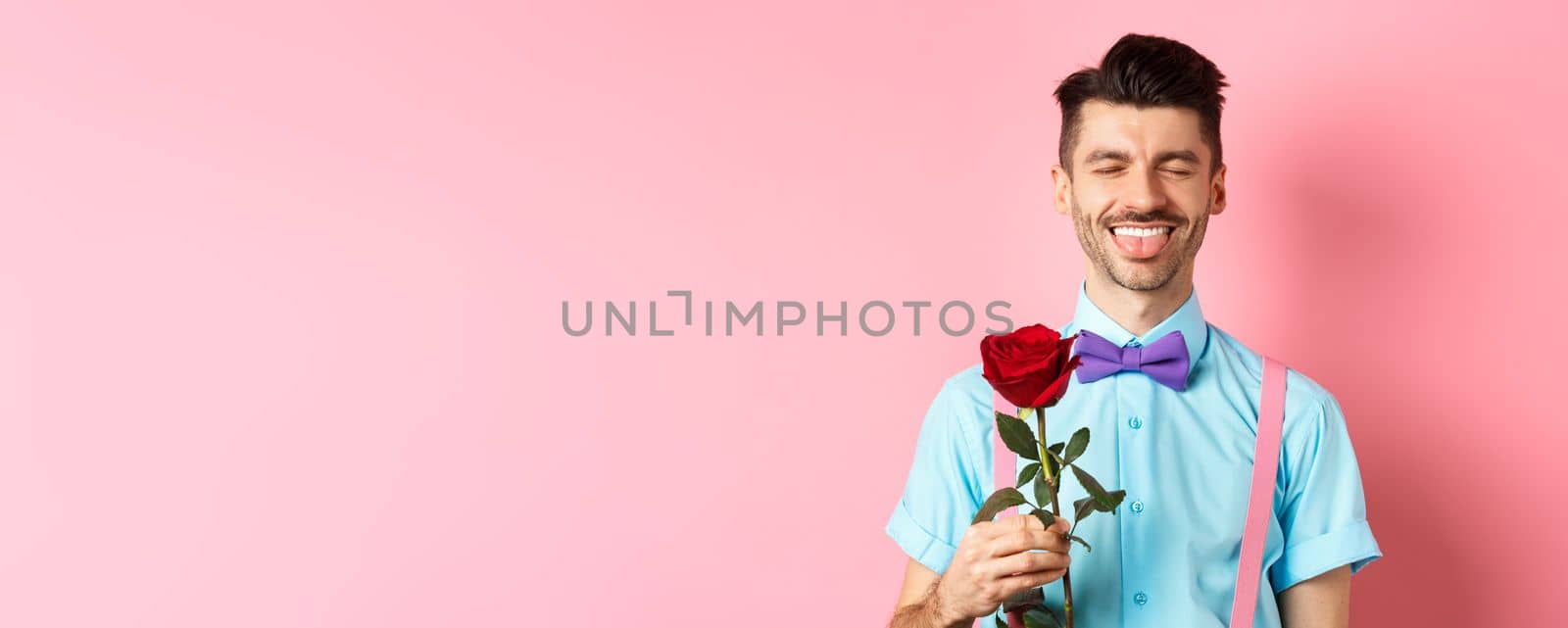 Happy man showing tongue and smiling, holding red rose for girlfriend on Valentines day, enjoying romantic date, pink background.