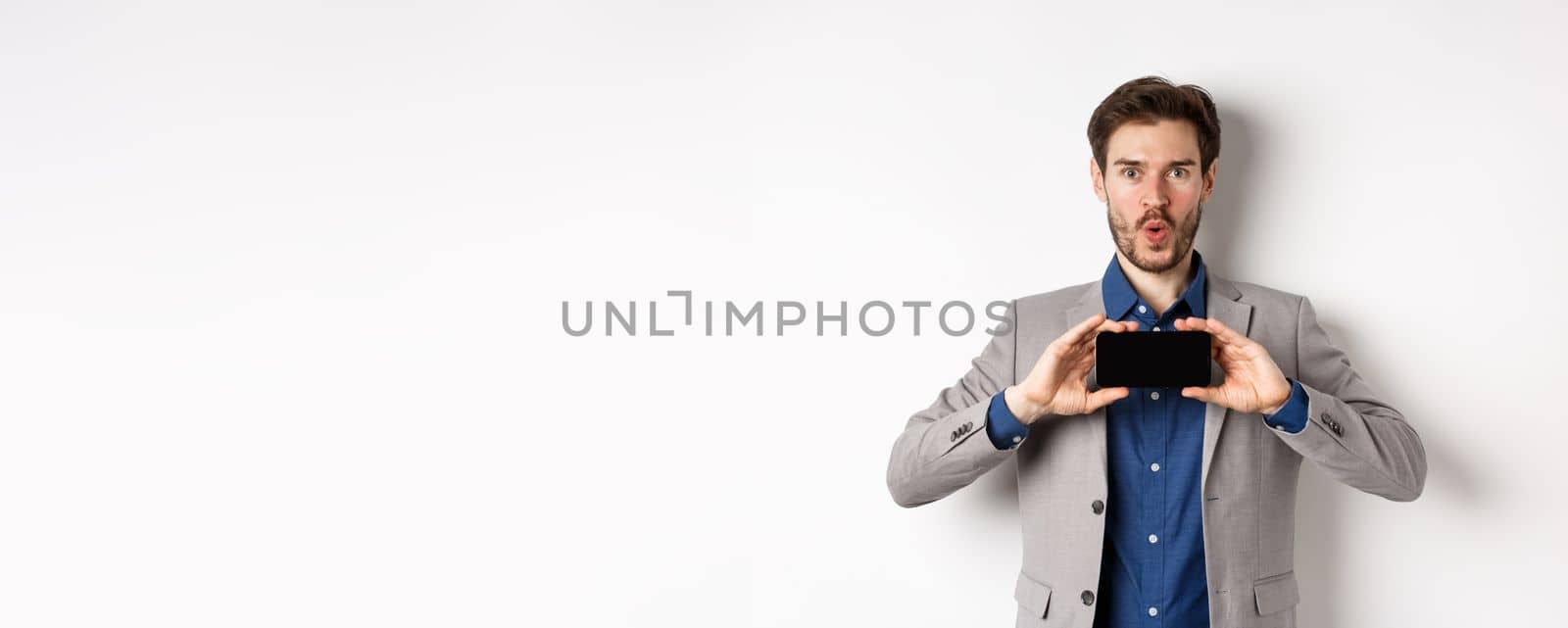 E-commerce and online shopping concept. Excited young man in business suit show empty smartphone screen and say wow amazed, white background.
