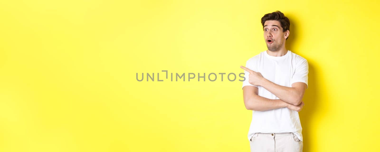 Impressed man in white t-shirt, looking and pointing finger left at promo, check out advertisement, standing against yellow background by Benzoix
