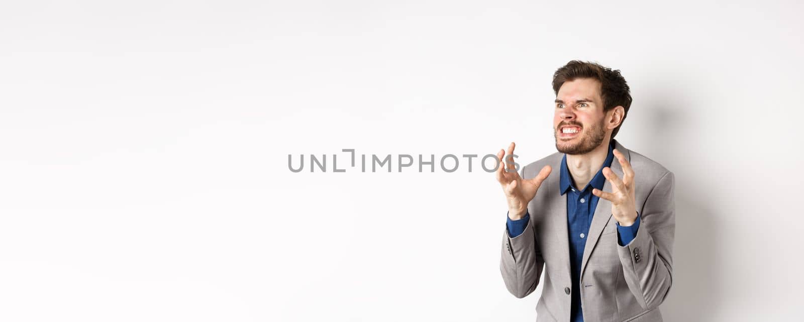 Angry man in business suit look left with hatred, stare aside and shaking hands with clenched teeth, standing mad and furious, wanting to kill someone, white background.