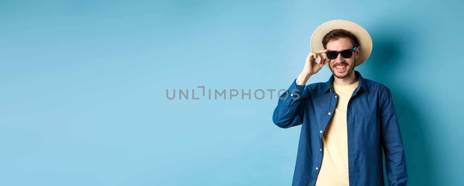 Happy smiling guy going on summer vacation, wearing straw hat and black sunglasses, standing on blue background.