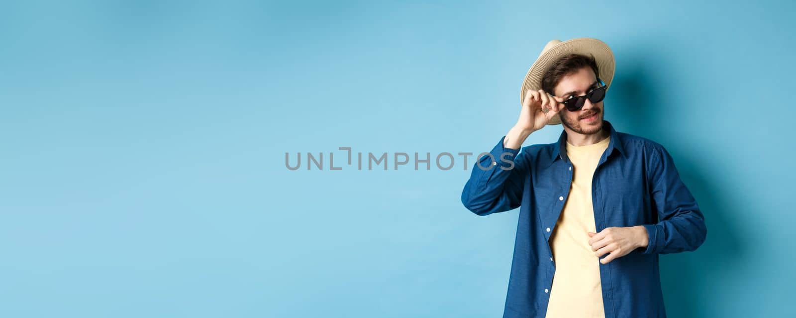 Handsome tourist in sunglasses and straw hat looking aside, smiling pleased, exploring sighsteeing on vacation, blue background by Benzoix