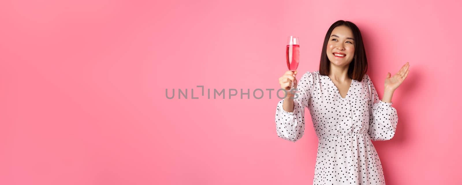 Beautiful asian woman, raising glass of champagne and smiling happy, drinking wine and celebrating, standing over pink background by Benzoix
