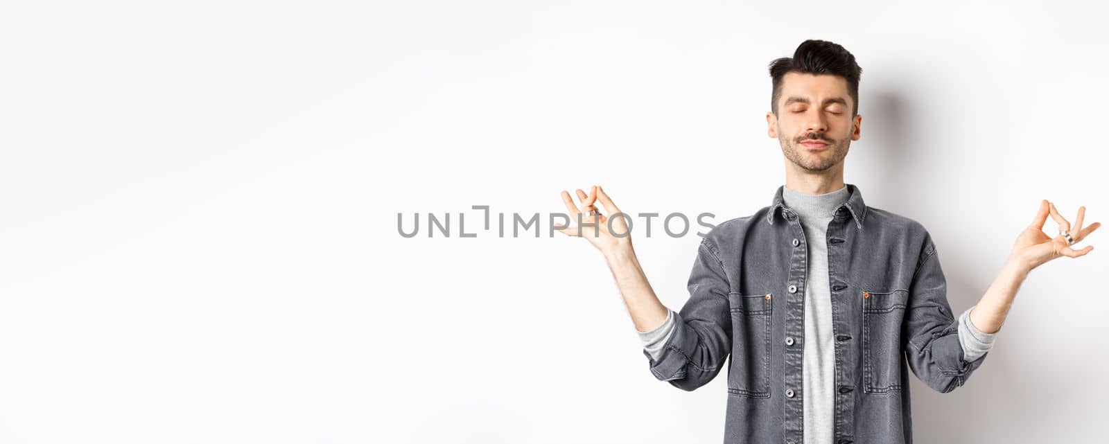 Calm guy meditating with peaceful face, holding hands in zen mudra sign and close eyes, practice yoga, relaxing on white background.