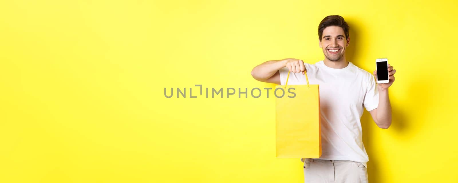 Concept of mobile banking and cashback. Young happy guy holding shopping bag and showing smartphone screen, yellow background.