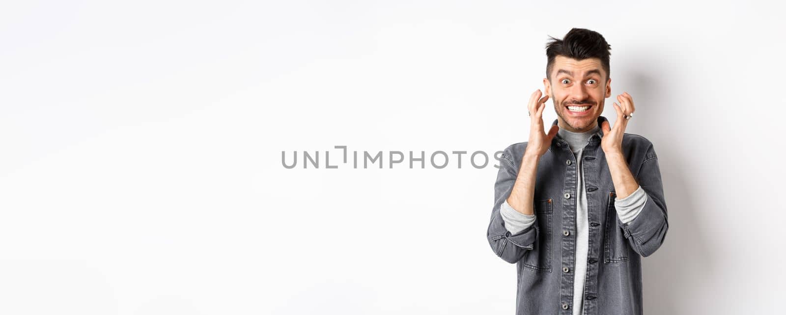 Angry man clench teeth and stare with hatred and anger at camera, trying to hold himself together, boiling from rage, standing on white background tensed.