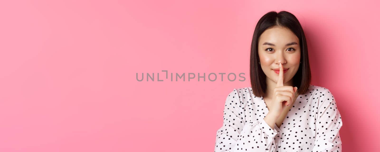 Close-up of mysterious asian woman hiding a secret, hushing and telling to keep quiet, standing over pink background.