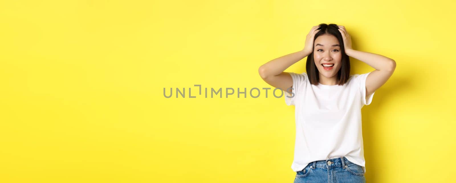 Amazed asian girl winning prize and looking with excitement and disbelief, holding hands on head and smiling happy, standing over yellow background.