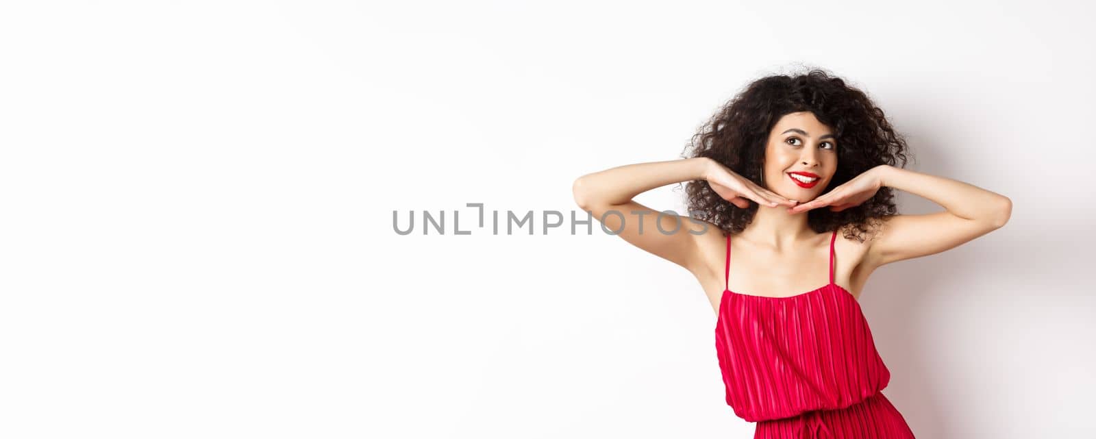 Beautiful lady with curly hair, wearing red dress, showing hear face with makeup and smiling, feeling carefree on white background.