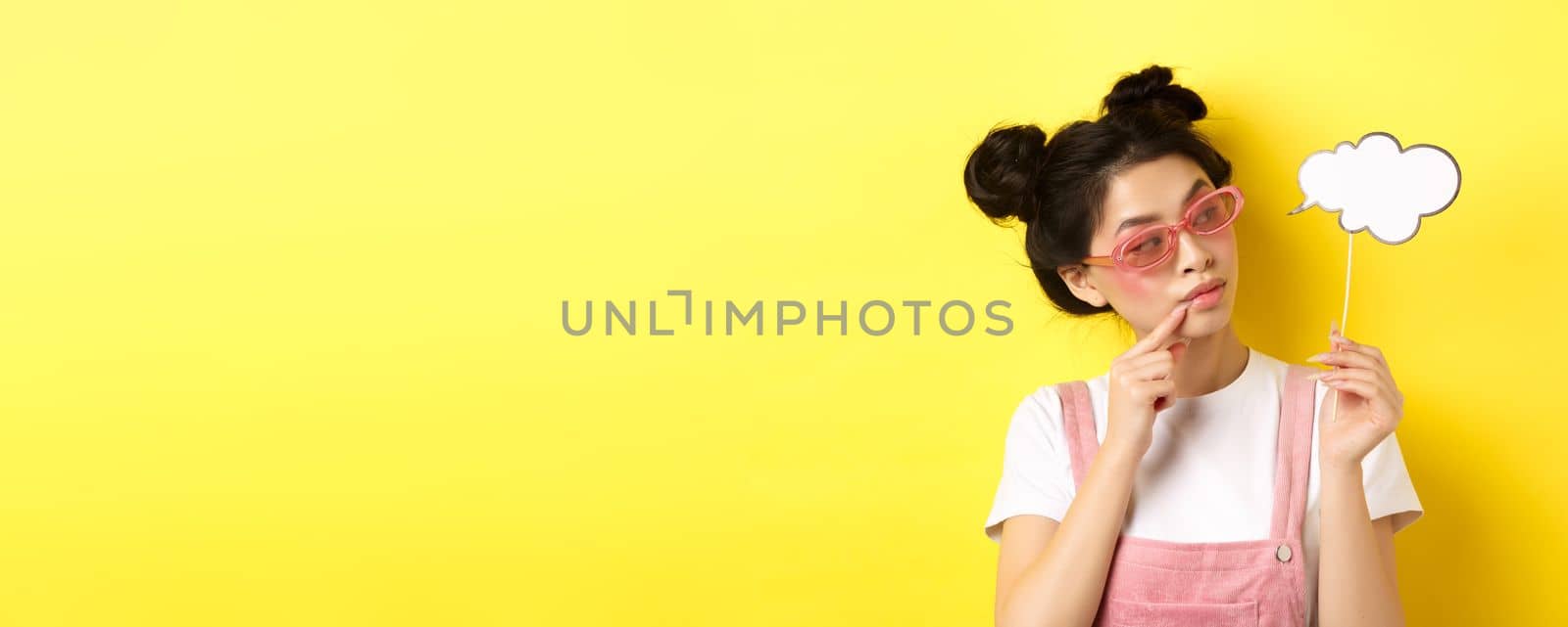 Summer and fashion concept. Stylish glamour asian girl in sunglasses, holding comment cloud party mask and looking aside, yellow background.