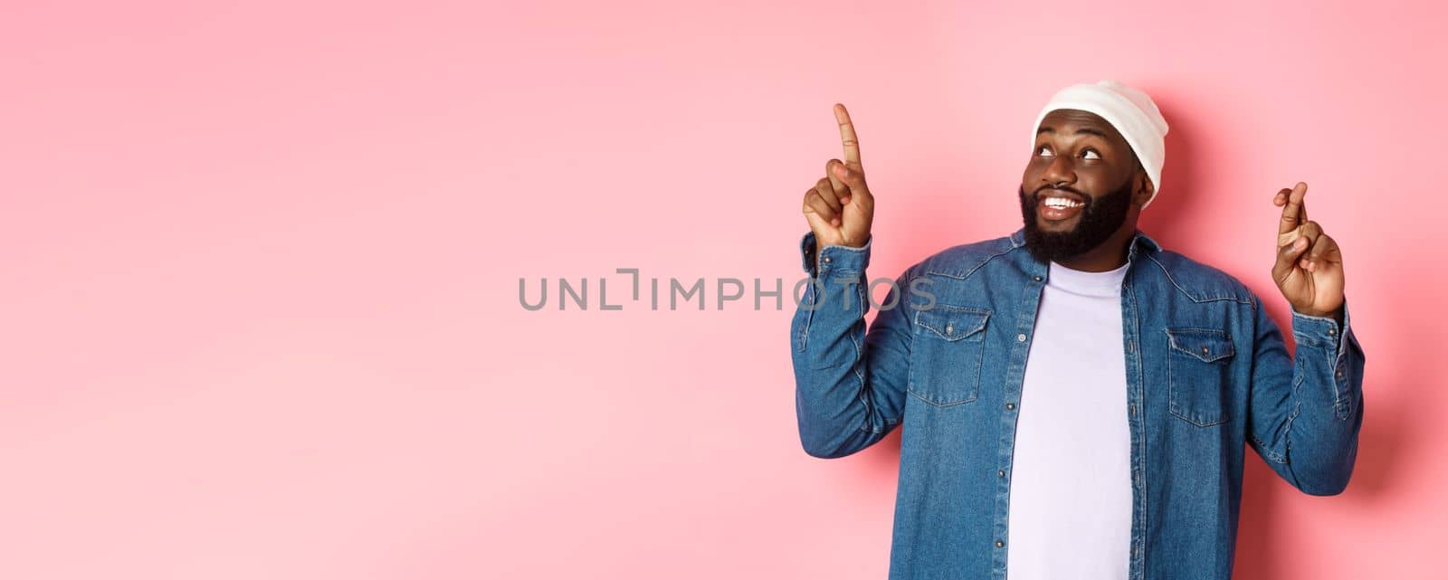 Hopeful african-american man making a wish, cross fingers for good luck, pointing and looking at upper left corner, dreaming about buying something, pink background.