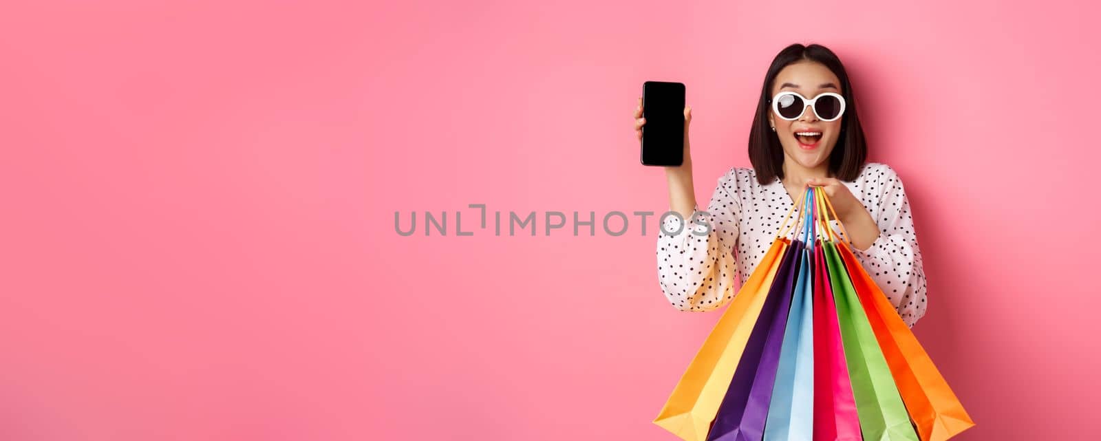 Attractive asian woman showing smartphone app and shopping bags, buying online via application, standing over pink background.