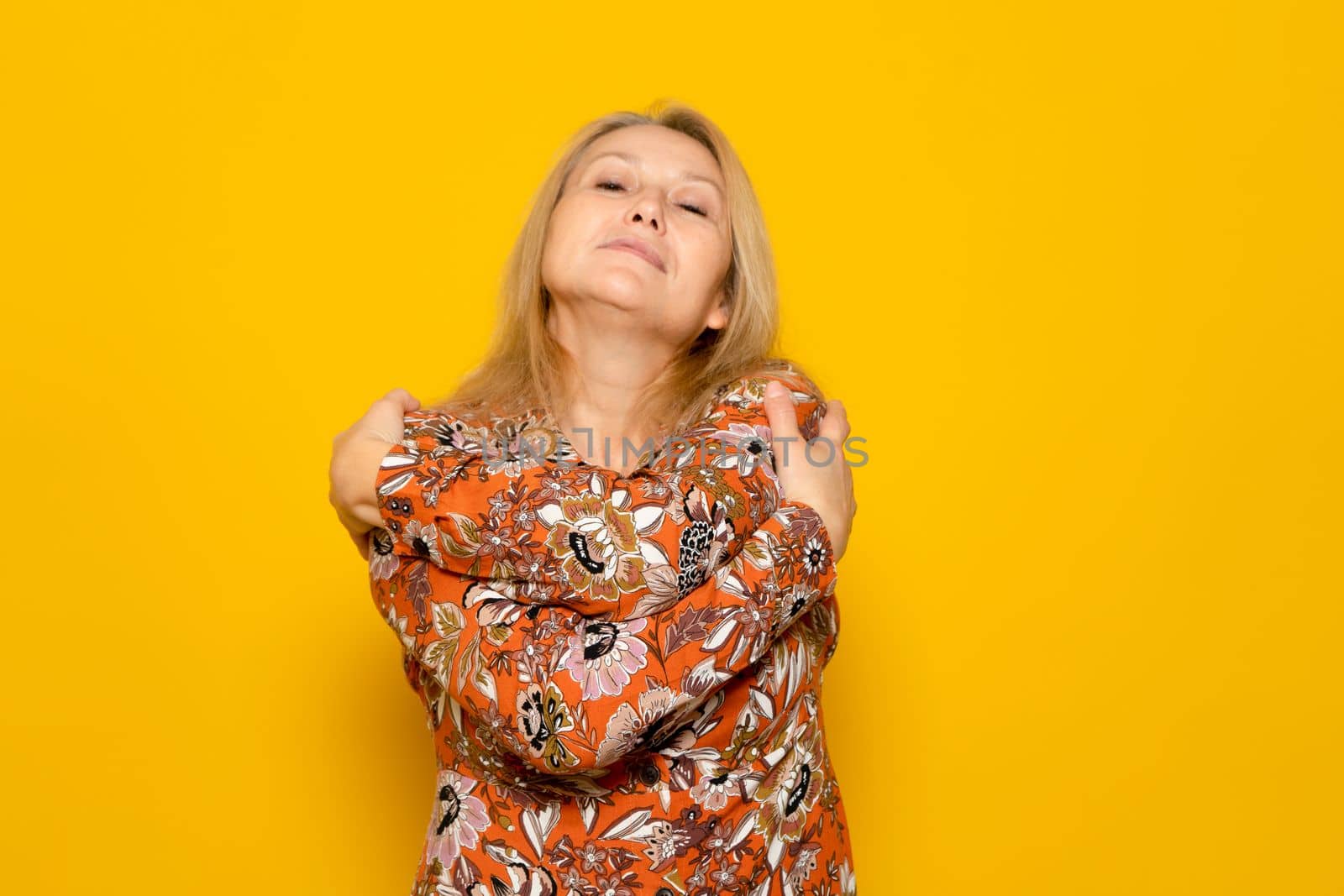 Close-up photo of a charming woman wearing a dress while smiling and hugging herself, isolated over yellow background