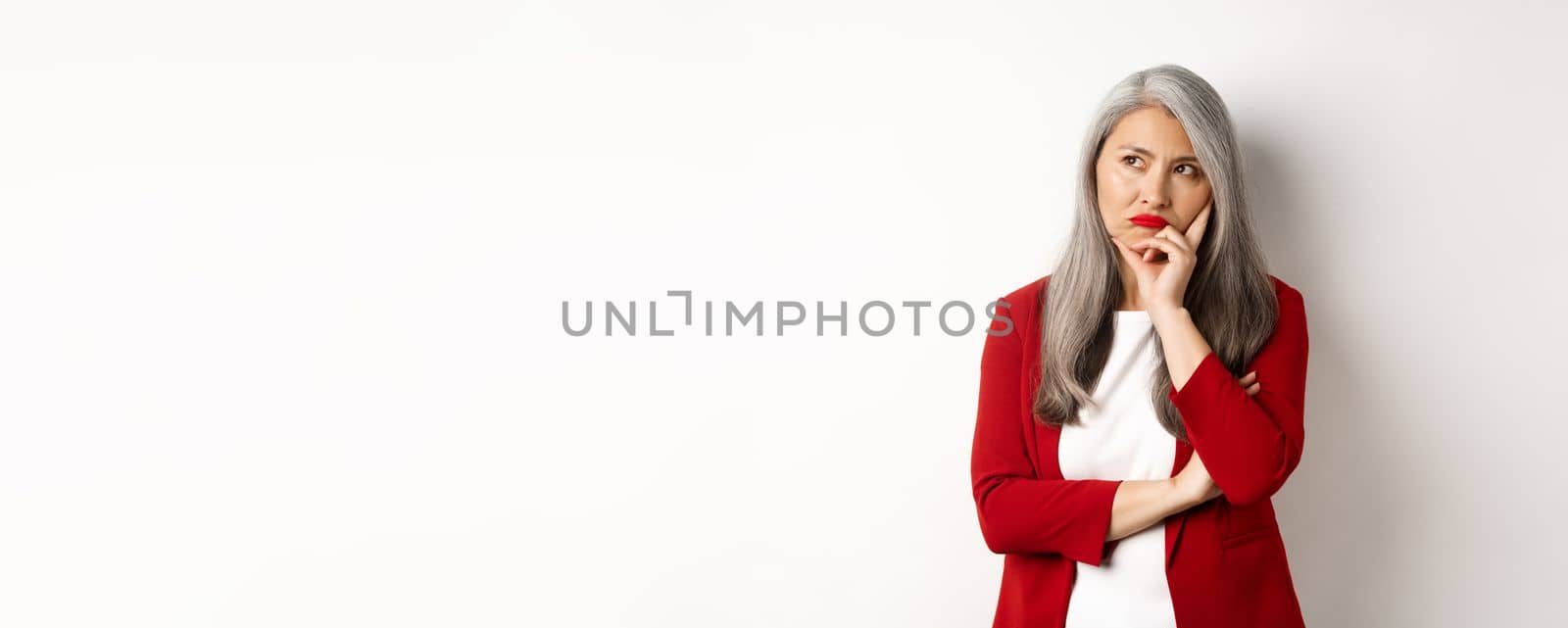 Troubled asian businesswoman in red blazer and lips, pouting and looking left annoyed, standing against white background by Benzoix