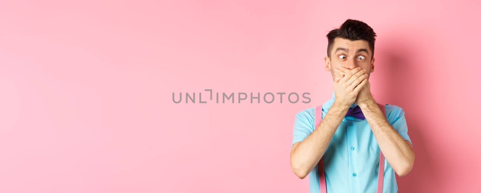 Funny young man covering mouth with hands and squint eyes, making clown grimaces, standing silly on pink background, entertain people.