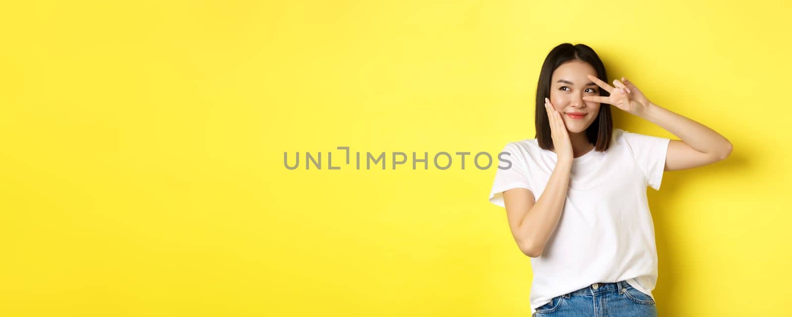 Lovely asian girl in white t-shirt posing with hand on cheek, showing peace sign on eye, standing over yellow background.