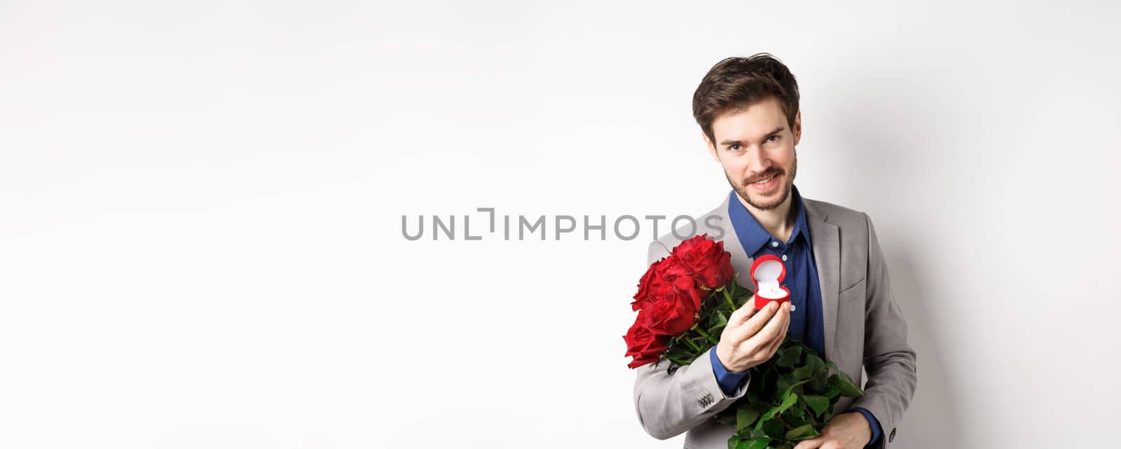 Romantic man with boquet of red roses asking to marry him, holding engagement ring and looking confident at camera, standing in suit over white background.