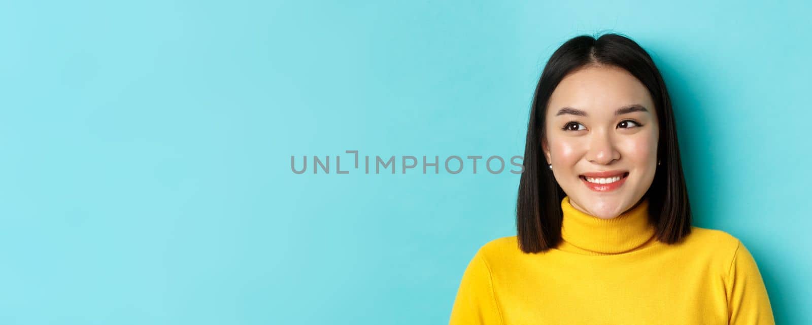 Beauty and makeup concept. Dreamy asian girl with perfect healthy skin, looking left and smiling happy, standing over blue background by Benzoix