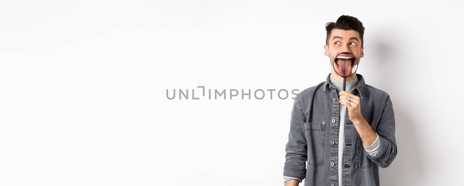 Handsome positive guy showing white perfect teeth and tongue with magnifying glass, looking left at logo, standing against white background by Benzoix