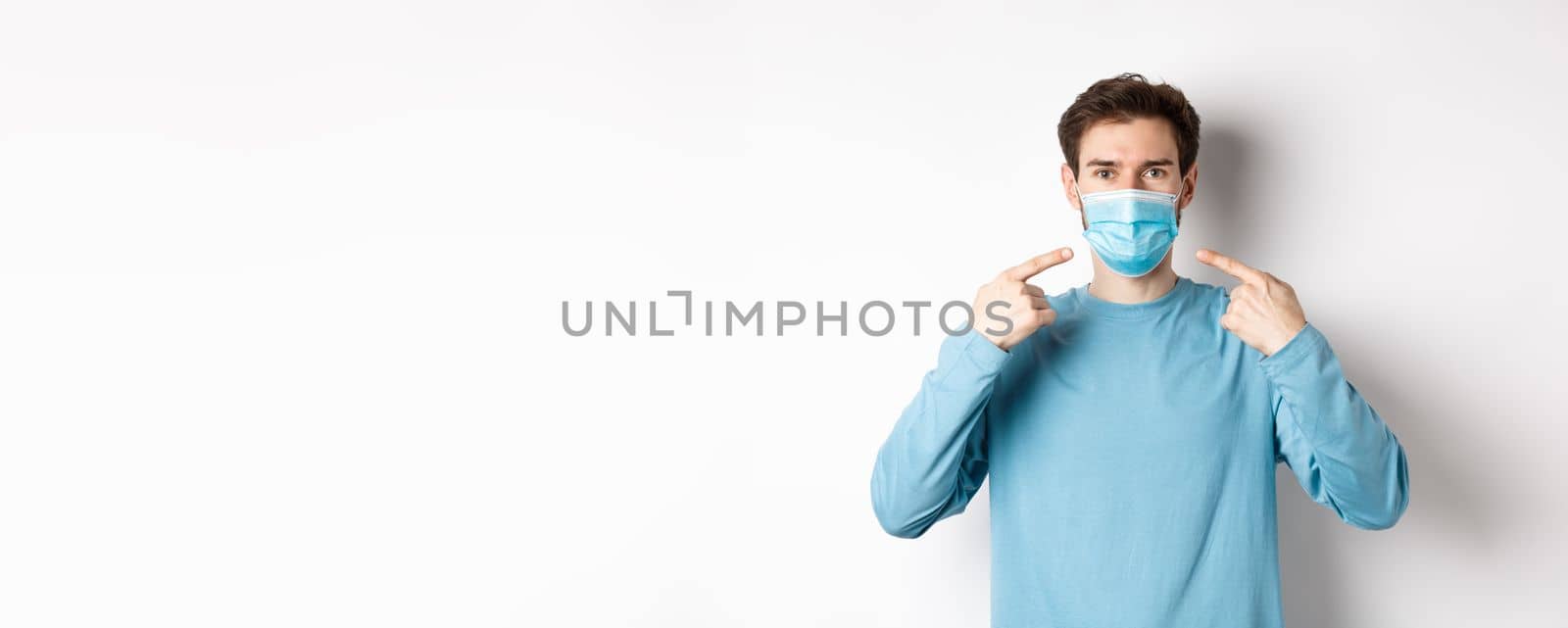 Coronavirus, health and quarantine concept. Young caucasian man in casual clothes pointing at his medical mask, asking to use preventive measures during pandemic, white background by Benzoix