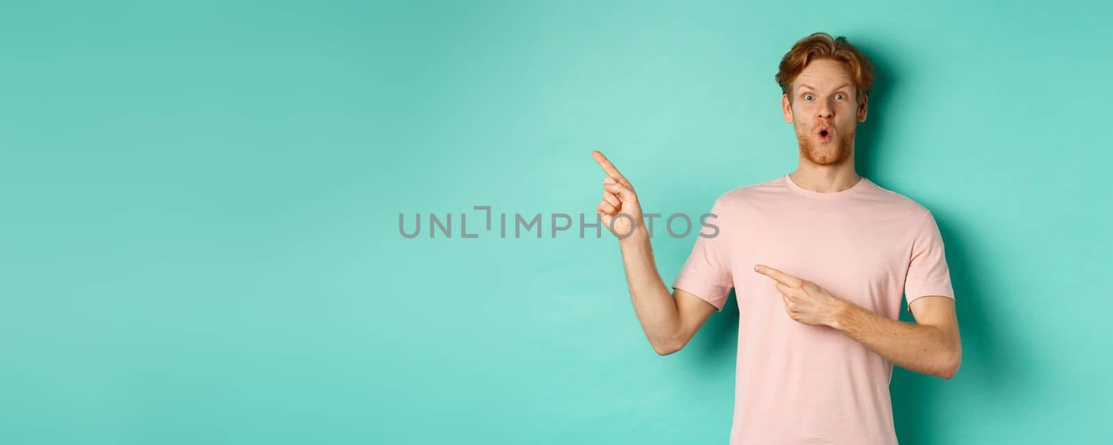 Amazed young man with red hair and beard, gasping amazed, pointing at upper left corner and showing promo, checking out advertisement, standing over turquoise background.