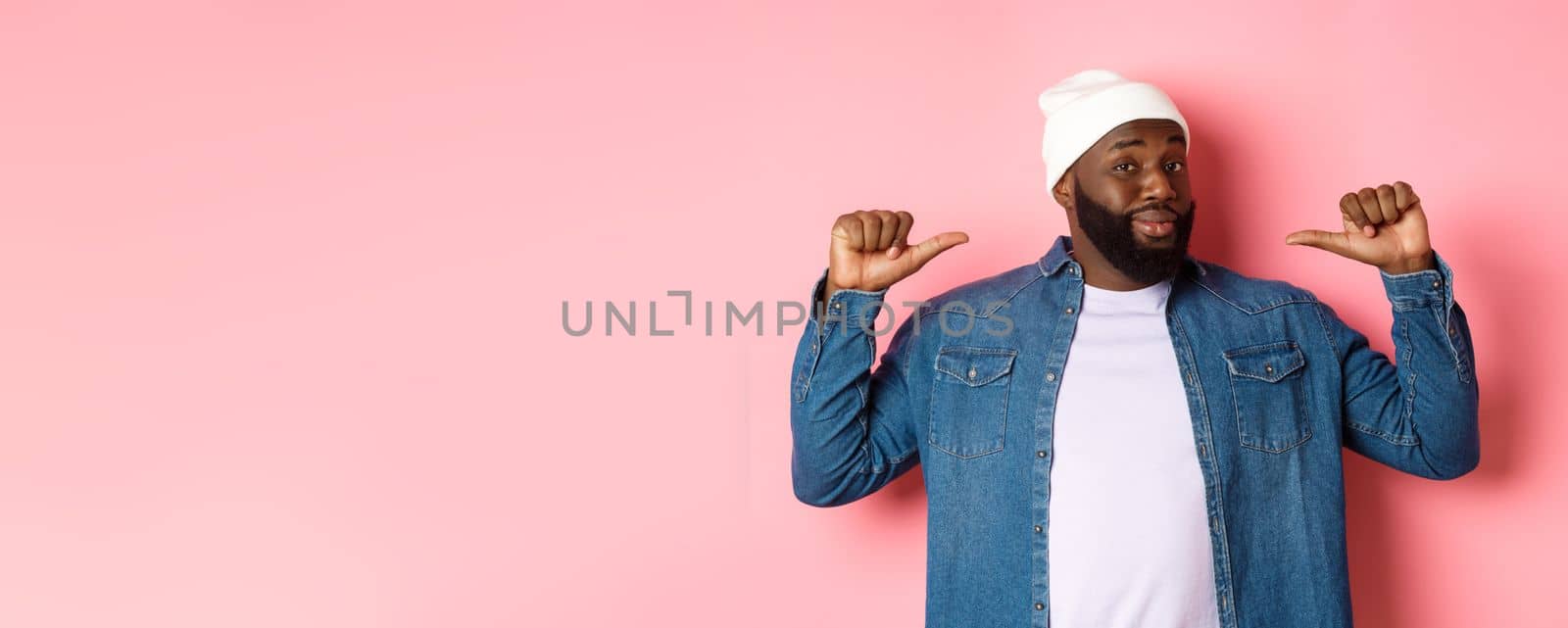 Confident and cheeky Black man pointing at himself, staring at camera self-assured, show-off, standing over pink background by Benzoix