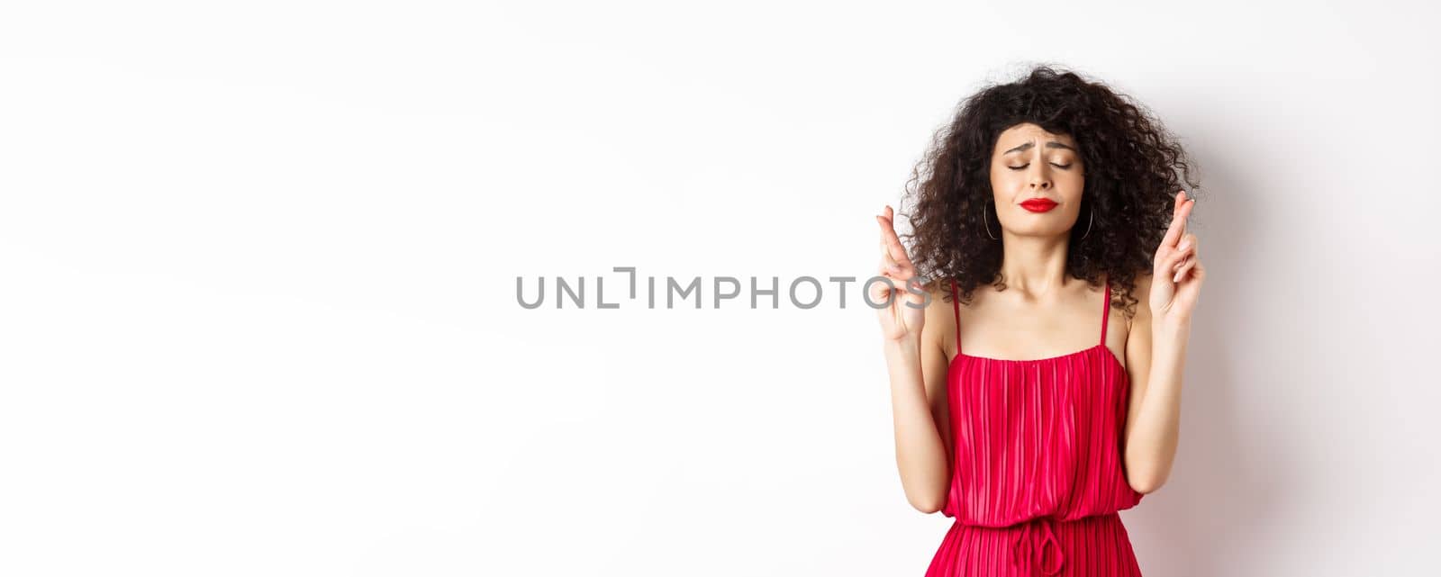 Hopeful woman in red dress making wish, praying with fingers crossed and closed eyes, pleading god, standing over white background.
