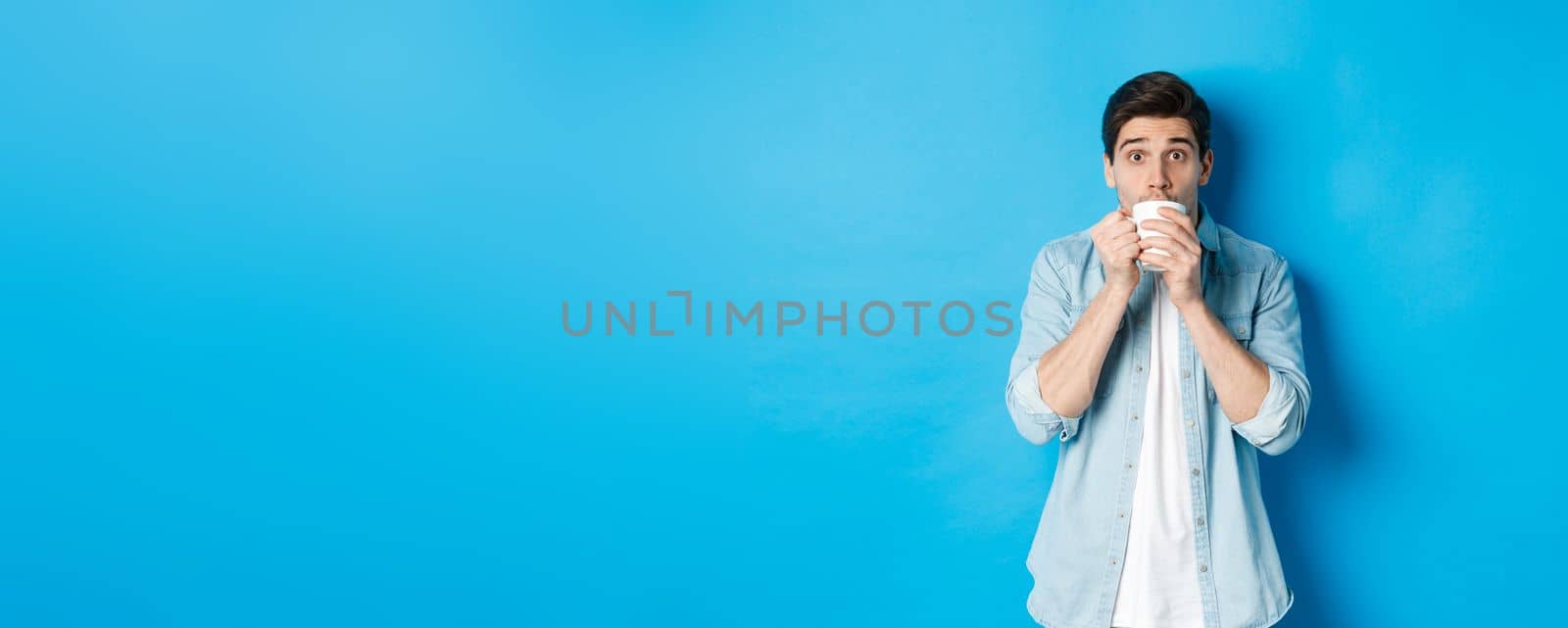 Man looking excited and sipping tea or coffee from white mug, standing over blue background by Benzoix