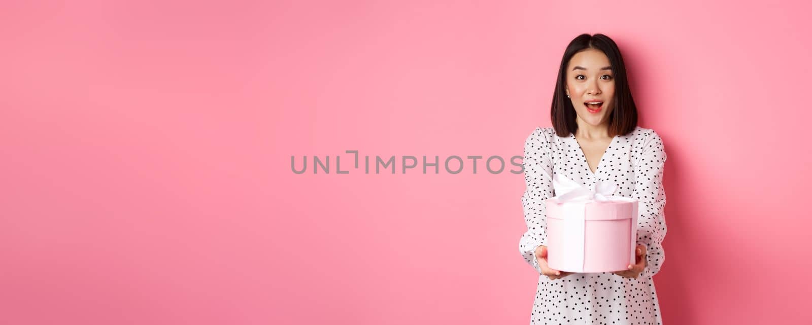 Cute asian woman congratulate with holiday or birthday, giving gift in cute box, standing over pink background.