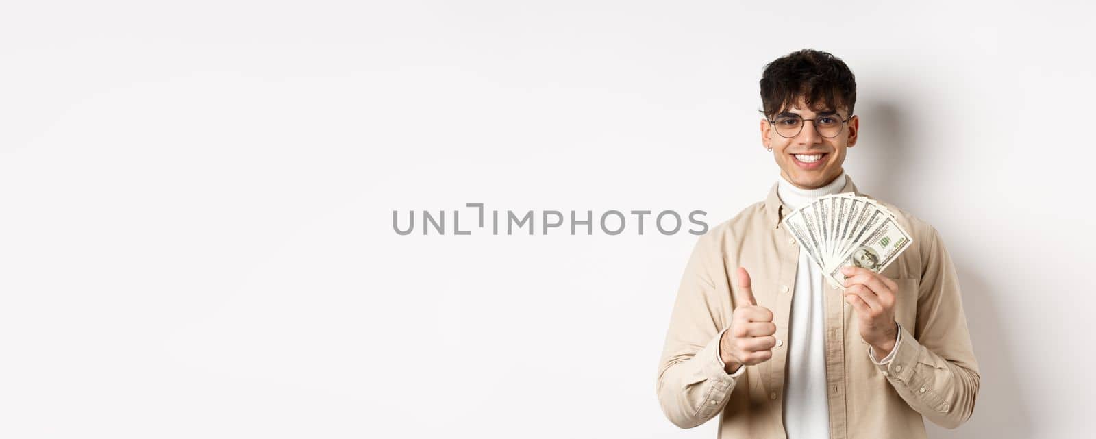 Young guy in glasses showing dollar bills and thumbs up, making money, standing with cash on white background.