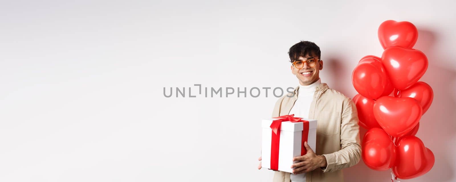 Valentines day and romance concept. Man in love prepare surprise gift for lover, holding present in box and standing near red hearts gesture, white background.
