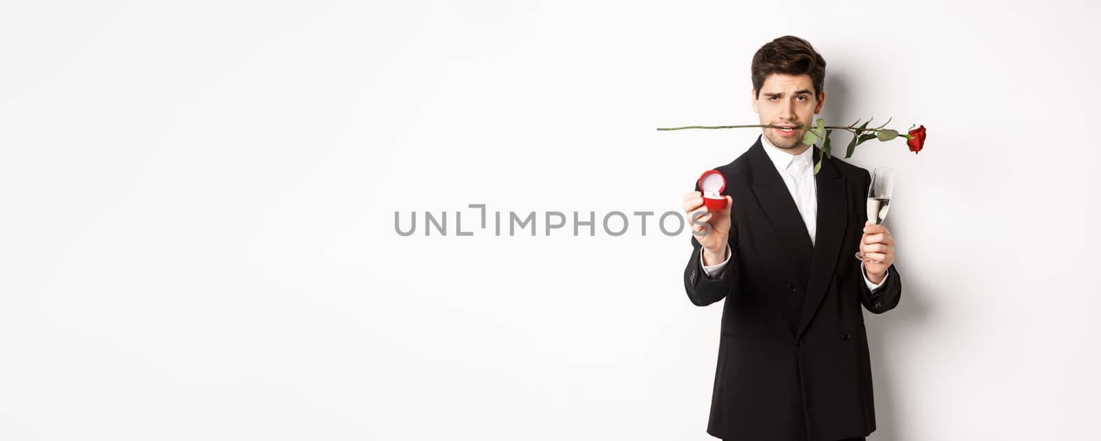 Passionate young man in suit making a proposal, holding rose in teeth and glass of champagne, showing engagement ring, asking to marry him, standing against white background.