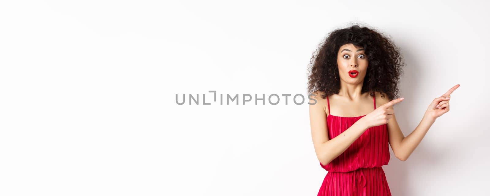 Surprised woman in red dress and makeup pointing fingers right, showing logo and look intrigued, standing on white background.
