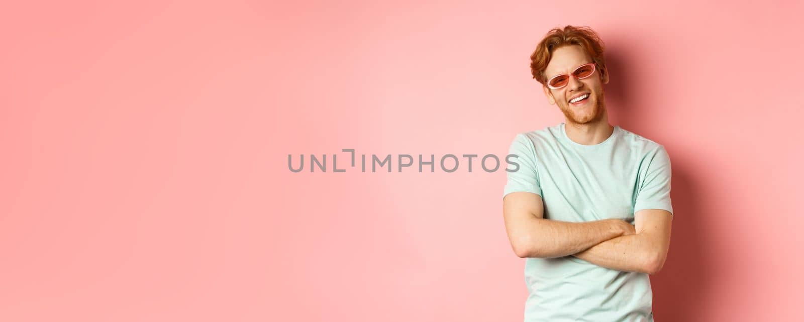 Tourism and vacation concept. Friendly young man with red hair, wearing sunglasses and t-shirt, cross arms on chest and smiling joyfully at camera, pink background by Benzoix