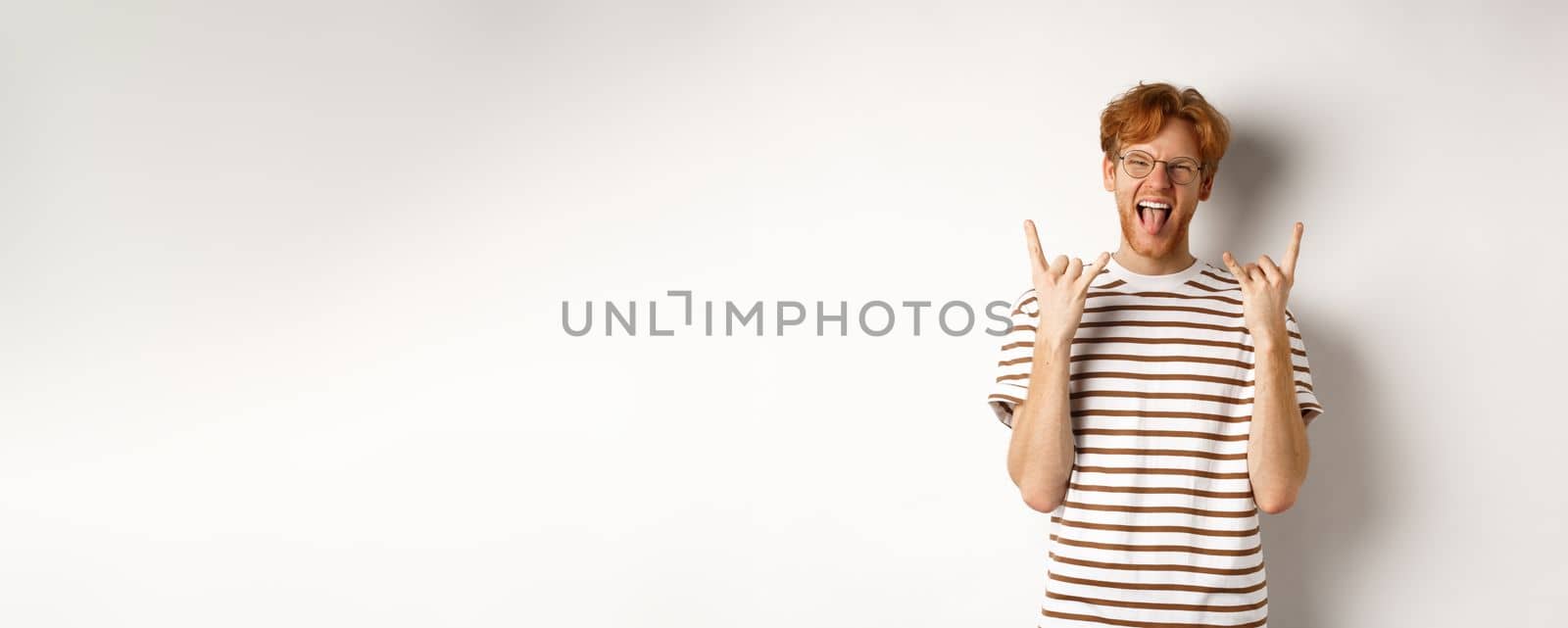Funny and happy redhead man having fun, showing rock-n-roll horn and sticking tongue, enjoying party, standing over white background by Benzoix