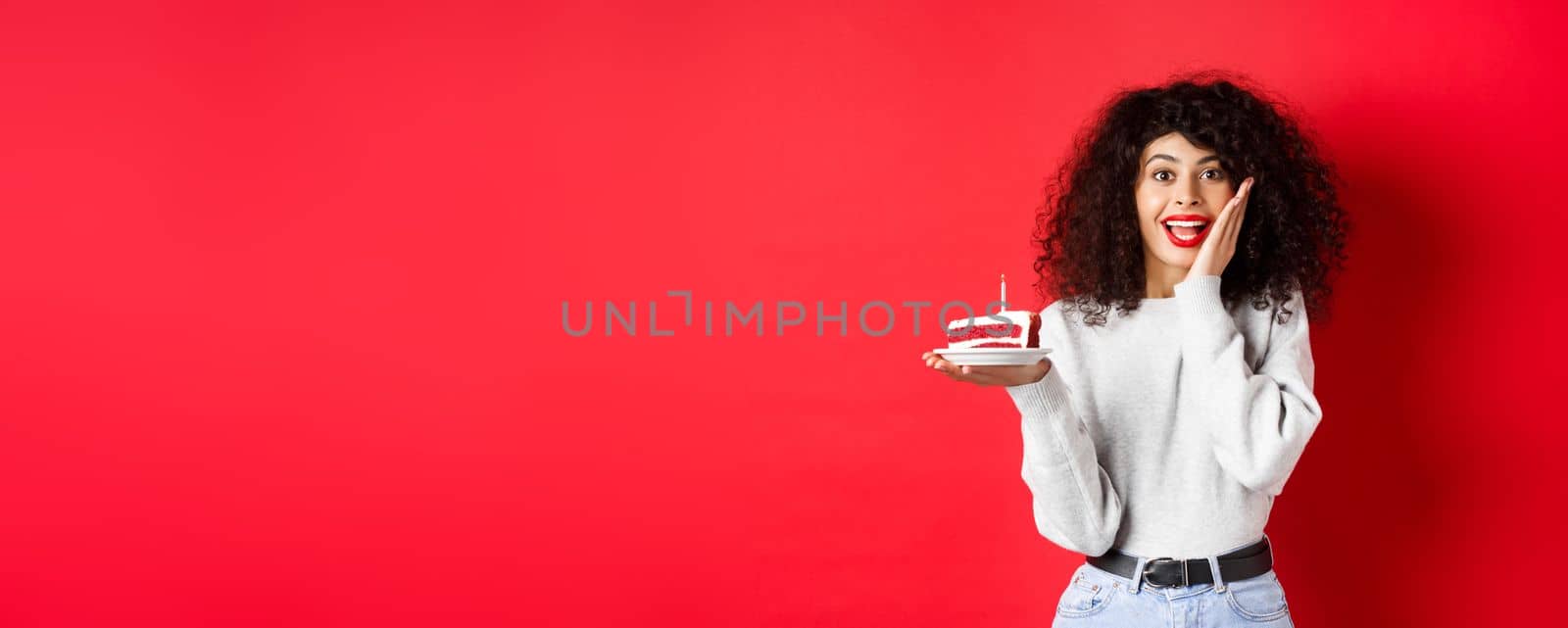 Excited woman celebrating birthday, holding cake and looking surprised and happy at camera, red background by Benzoix