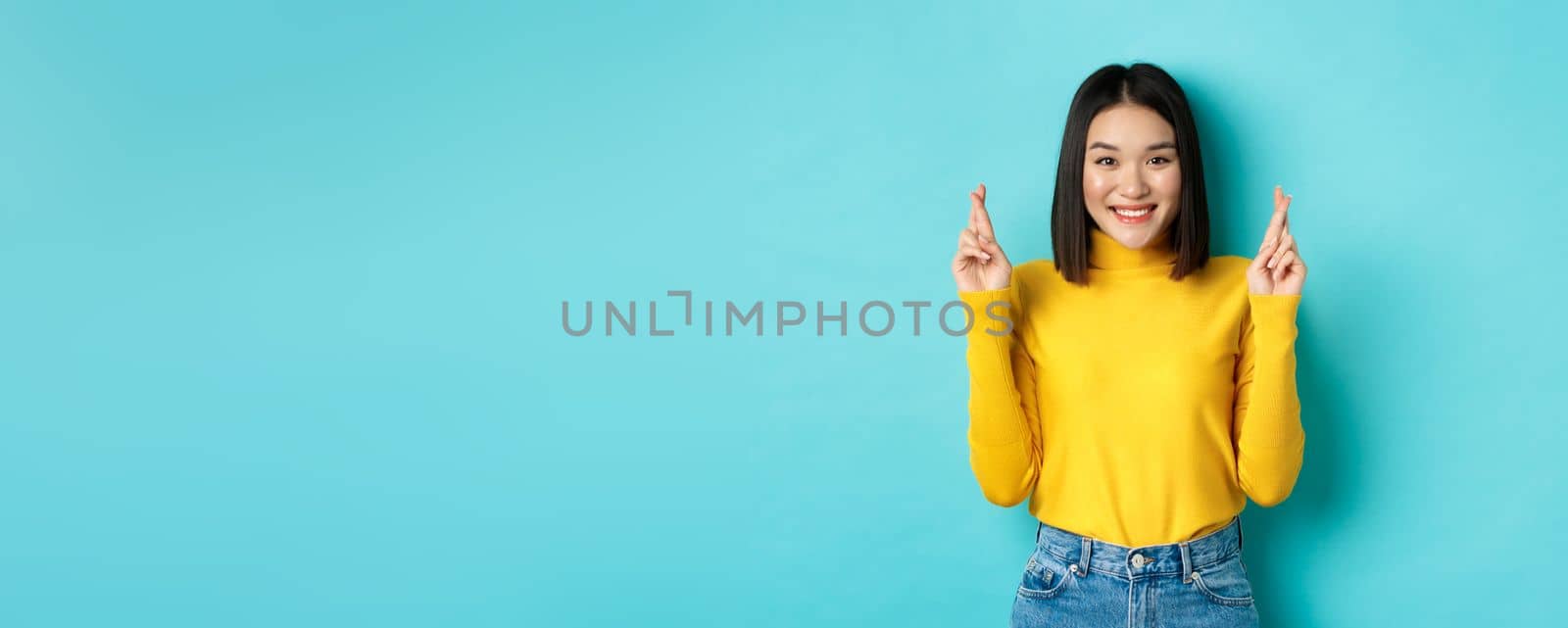 Hopeful and optimistic asian woman cross fingers for good luck, making wish and smiling, looking at camera and expecting something, standing over blue background by Benzoix