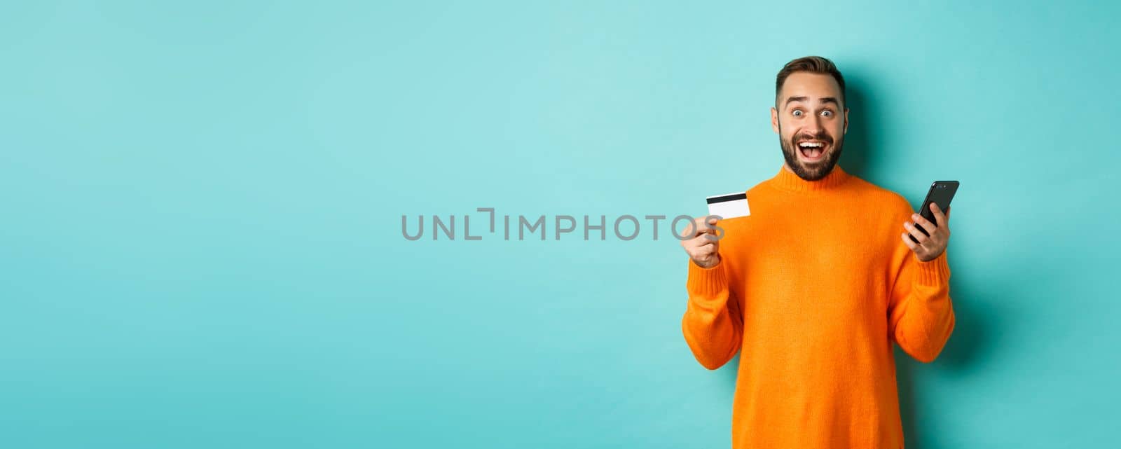 Online shopping. Surprised man holding mobile phone and credit card, paying in internet store, standing over light blue background.