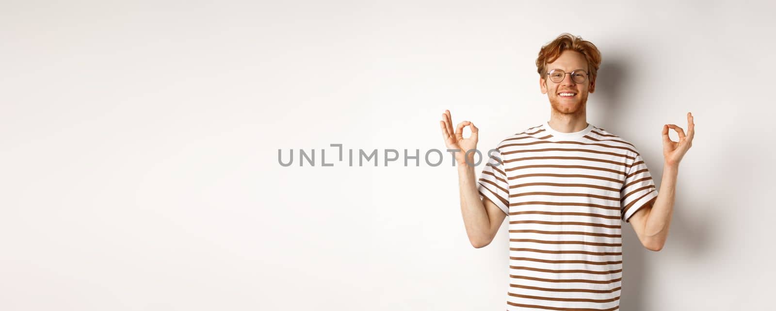 Cheerful redhead man in glasses have all under control, showing okay signs and smiling satisfied, standing relaxed over white background.