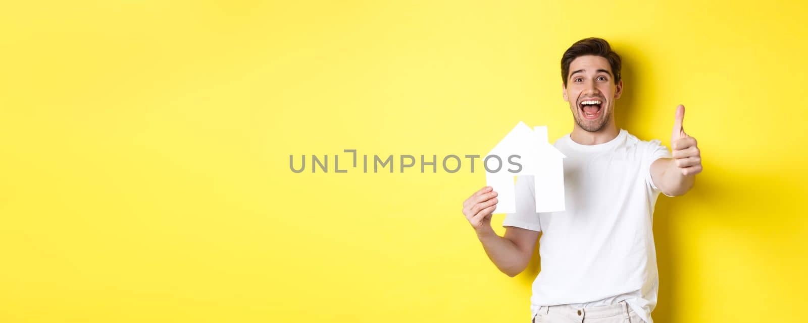 Real estate concept. Happy young male buyer showing thumb up and paper house model, smiling satisfied, standing over yellow background.