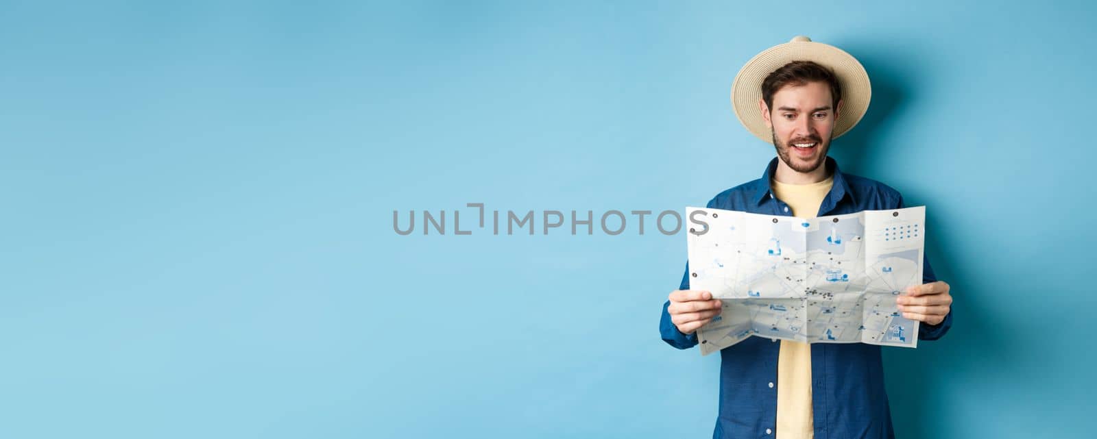 Cheerful tourist in summer hat planning travel route on vacation, looking at sighseeing map and smiling excited, standing on blue background by Benzoix