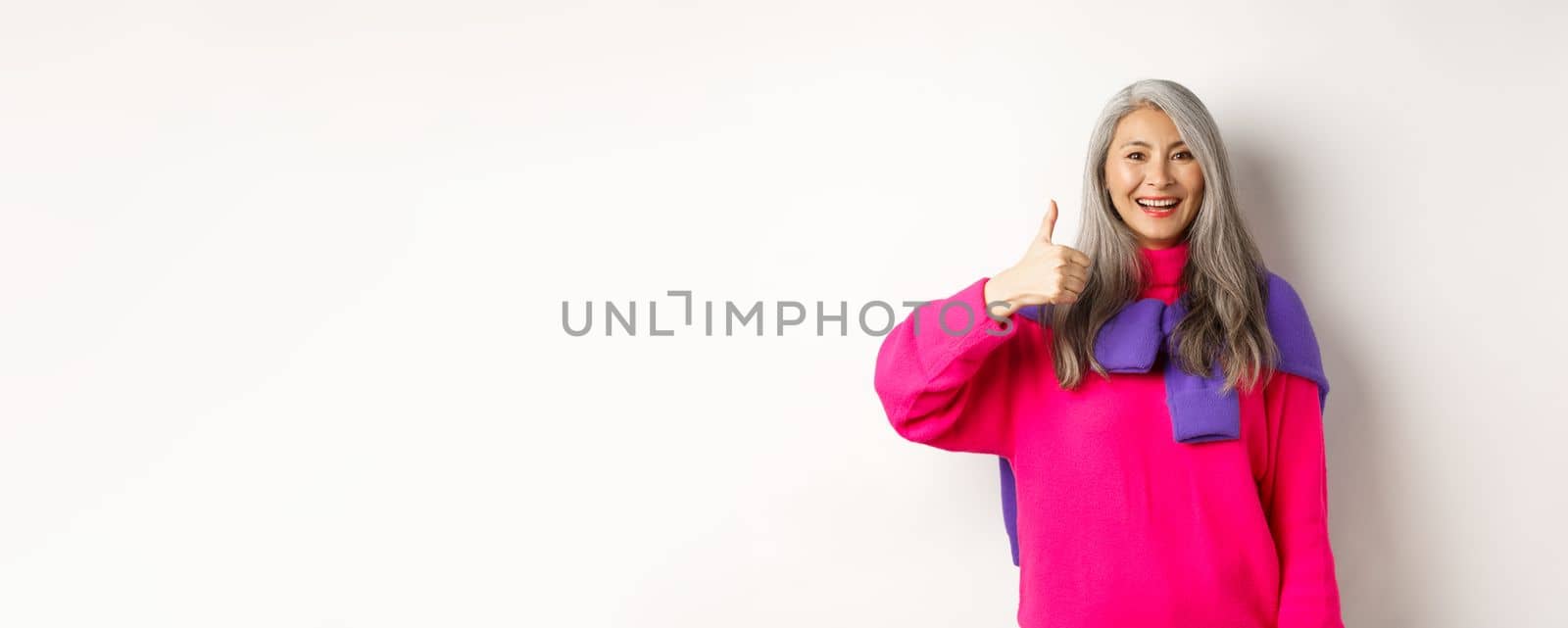 Portrait of happy and satisfied asian senior woman showing thumb-up, approval and like something, smiling and praising product, standing over white background by Benzoix