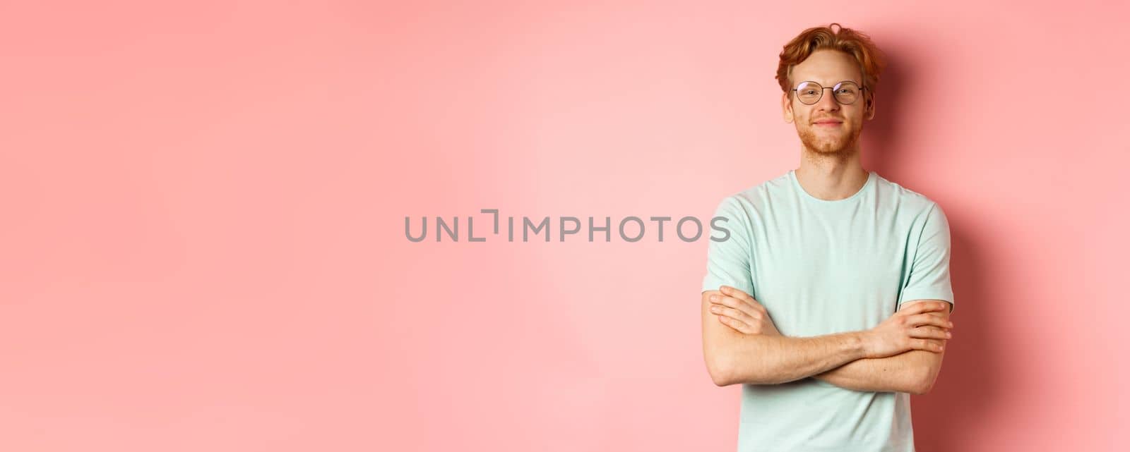 Portrait of satisfied caucasian man with red hair and beard, cross arms on chest and smiling with smug face, wearing glasses, standing over pink background.