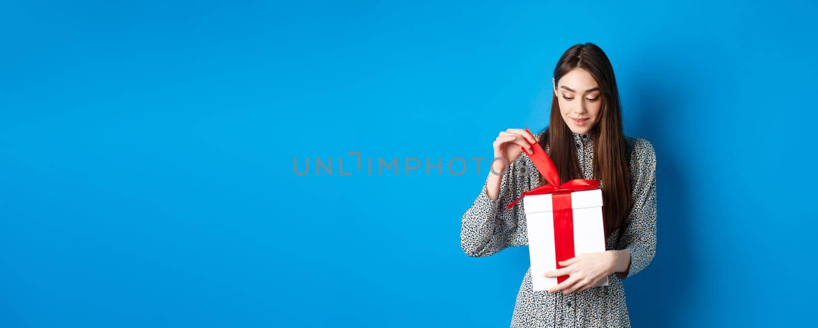 Valentines day. Cute young woman open box with gift, take-off ribbon from present and smiling intrigued, standing on blue background.