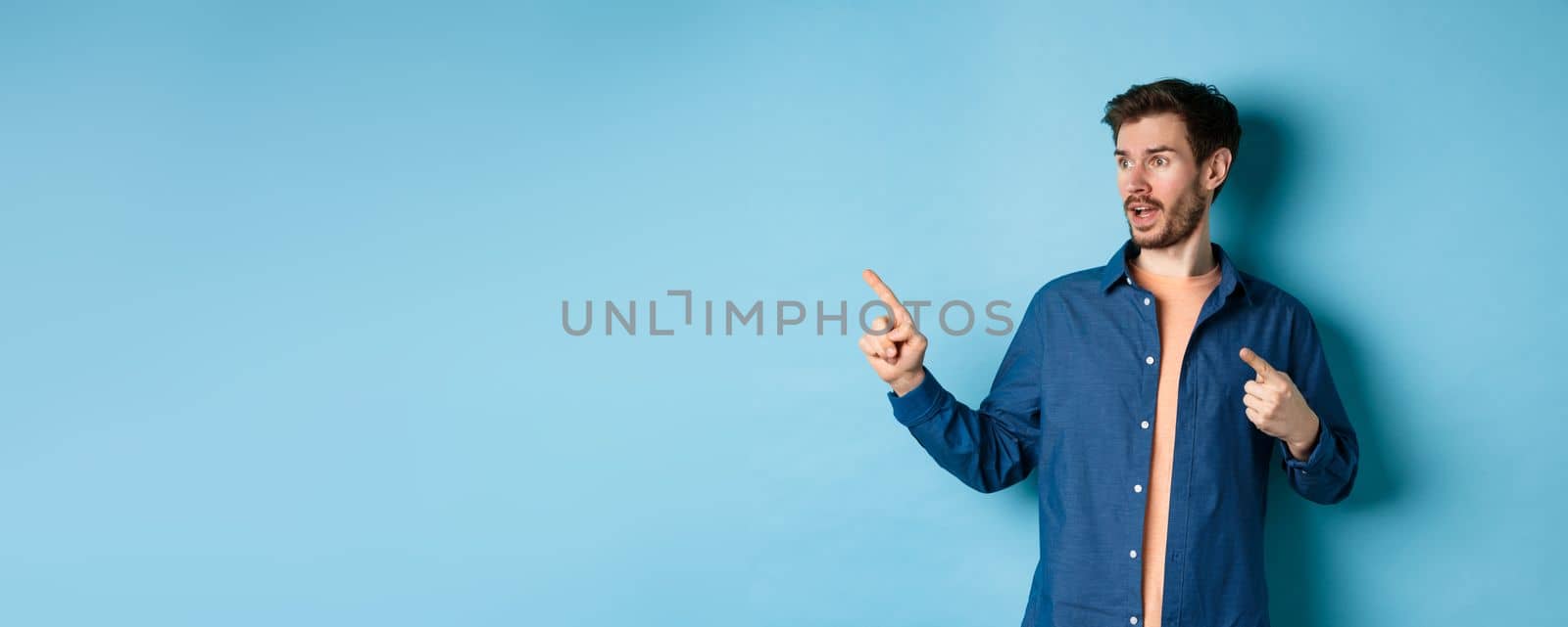 Amazed caucasian guy pointing and looking left, checking out advertisement, standing on blue background.
