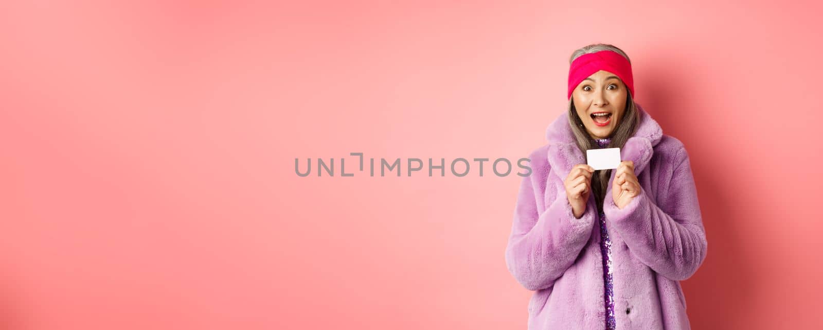 Shopping and fashion concept. Beautiful asian middle-aged woman showing plastic credit card and smiling amazed in stylish clothes, pink background.