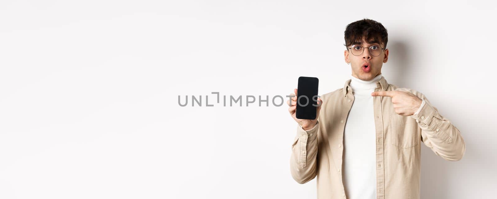 E-commerce concept. Portrait of young man pointing at mobile phone screen, showing advertisement online, standing on white background by Benzoix