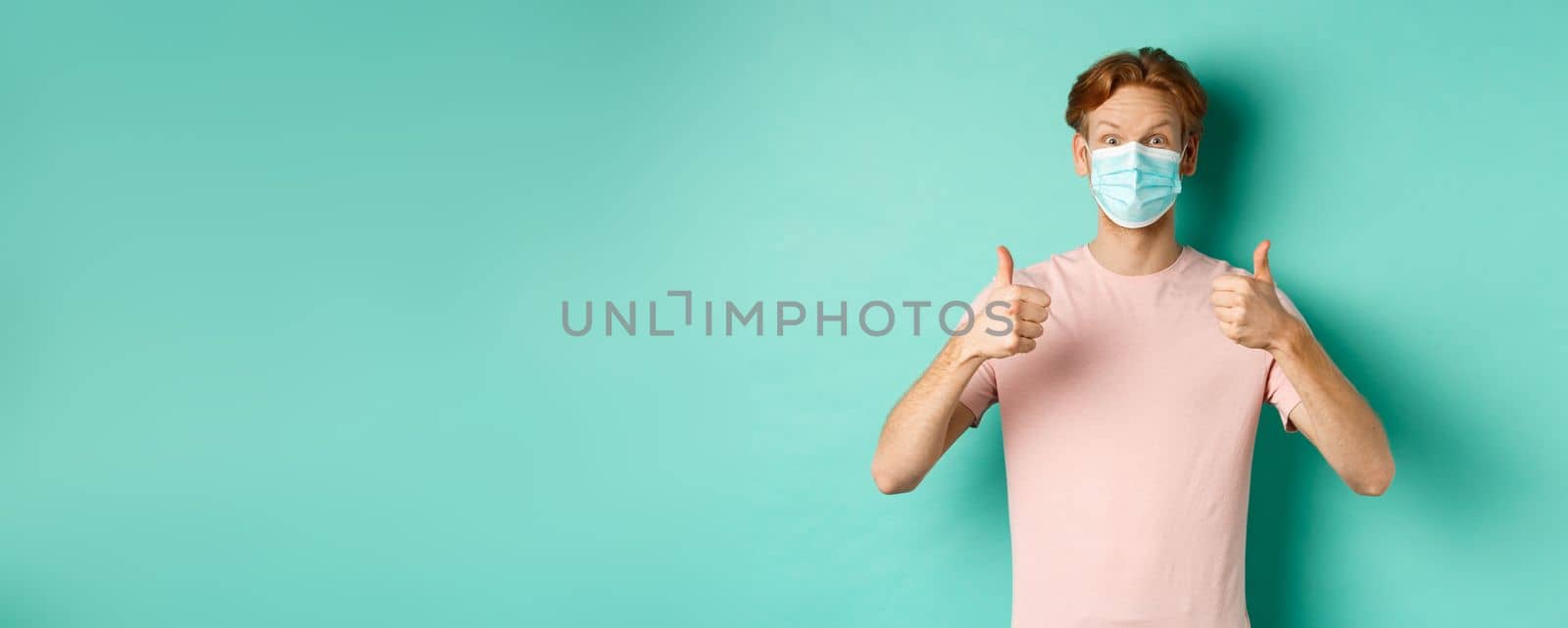 Covid-19, pandemic and lifestyle concept. Cheerful redhead guy in medical mask showing thumbs up in approval, like and praise product, standing over turquoise background.