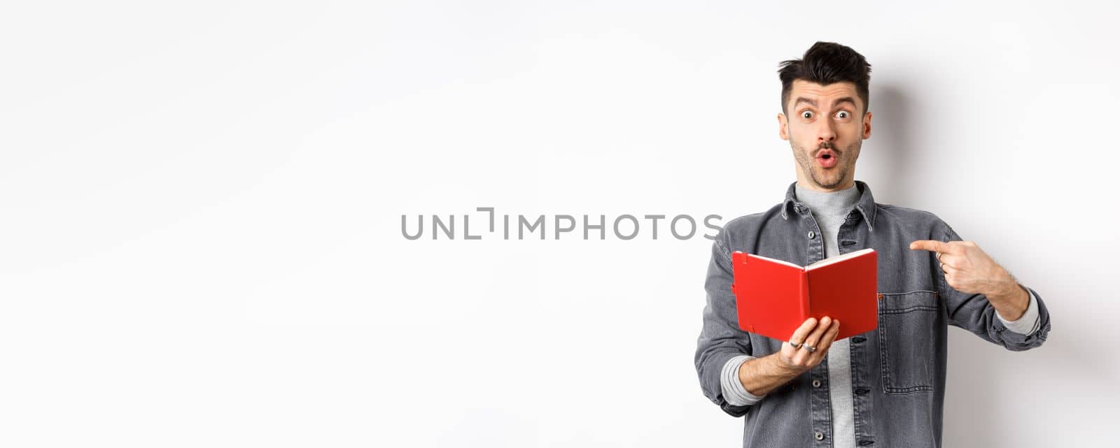 Excited guy read interesting thing in journal, pointing at planner and look impressed, standing against white background by Benzoix